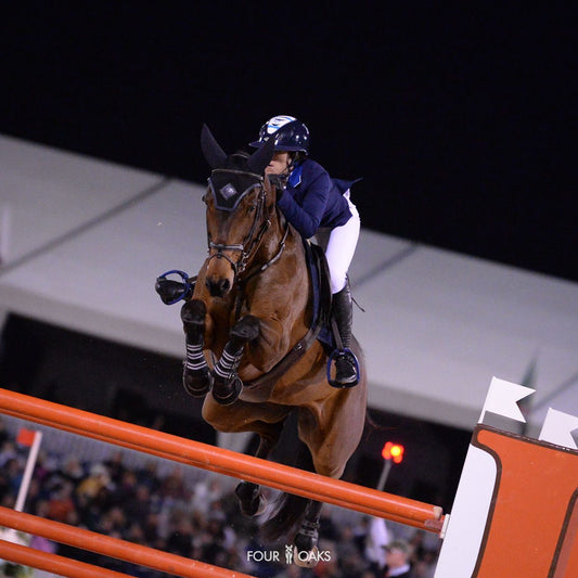 Sydney Shulman Desiderio competing in a night showjumping event, clearing an obstacle on a bay horse, showcasing professional equestrian skill